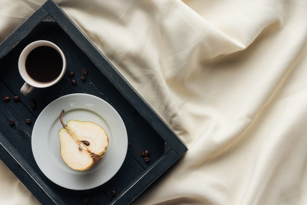 top view of cup of coffee with half of ripe pear on beige cloth, breakfast in bed concept